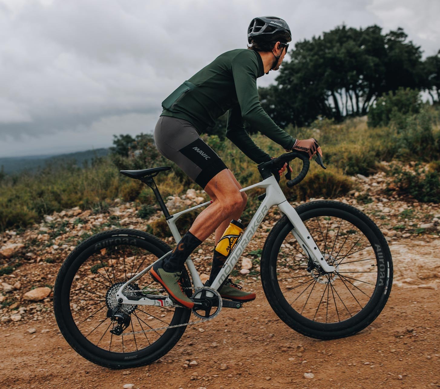 Ciclista de gravel con casco Mavic y ropa de ciclismo Mavic sobre una bicicleta gravel blanca en un camino pedregoso bajo cielo nublado