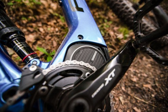 Detailfoto eines liegenden E-MTB mit blauem Rahmen. Man sieht die rechte Seite eines Shimano-Mittelantriebs und Teile von Kurbel und Kettenblatt. im Hintergrund unscharf Waldboden.