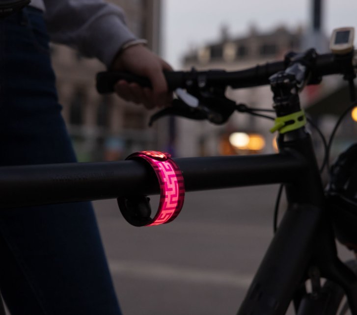 Ein pink leuchtendes Armband hängt am Oberrohr eines Fahrrads. In der Dämmerung ist eine urbane Szene erkennbar.