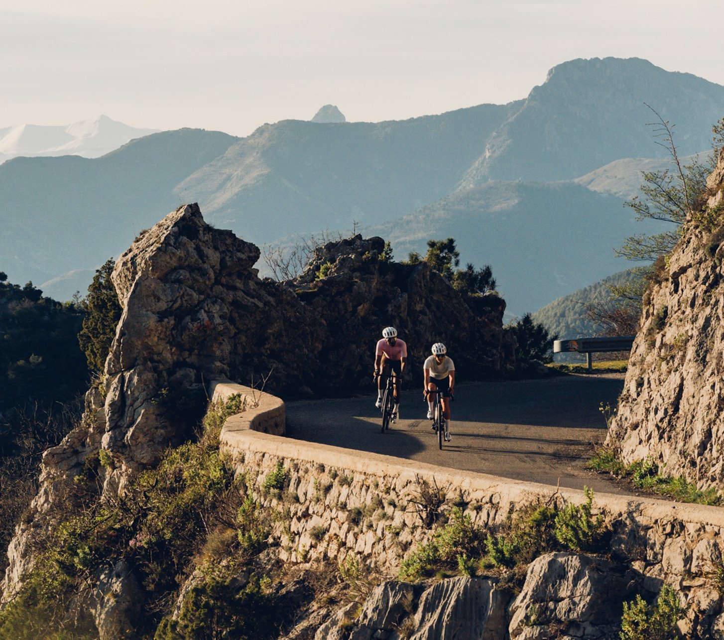 Zwei Rennradfahrer fahren auf kurviger Bergstraße zwischen Felsen durch die Alpenlandschaft – sportlicher Aufstieg in beeindruckender Kulisse bei tiefstehender Sonne