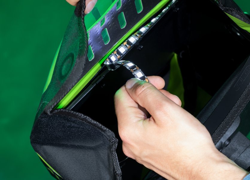 Eine Hand hält eine LED-Lichtleiste, die aus einer schwarzen Tasche mit grünen Akzenten herausgezogen wird. Die Tasche hat ein perforiertes Design an der Oberseite, durch das das Licht der LEDs scheint. Der Hintergrund ist dunkelgrün.