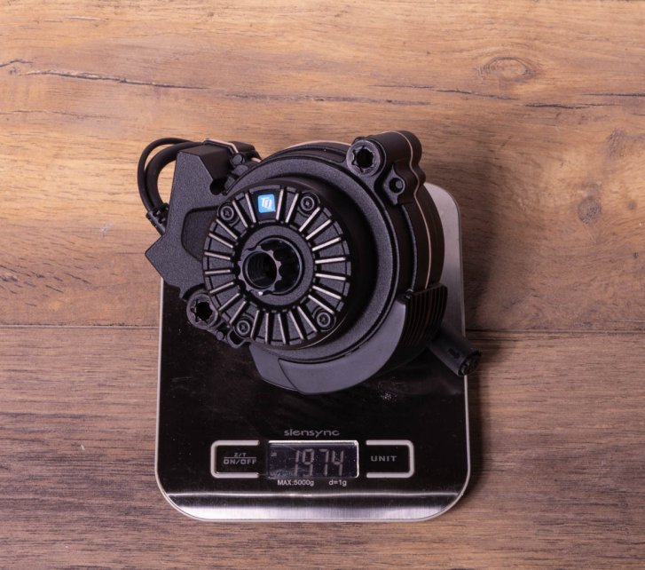 A TQ HPR60 e-bike drive on a kitchen scale on a dark wooden floor. Display shows 1,974 grams