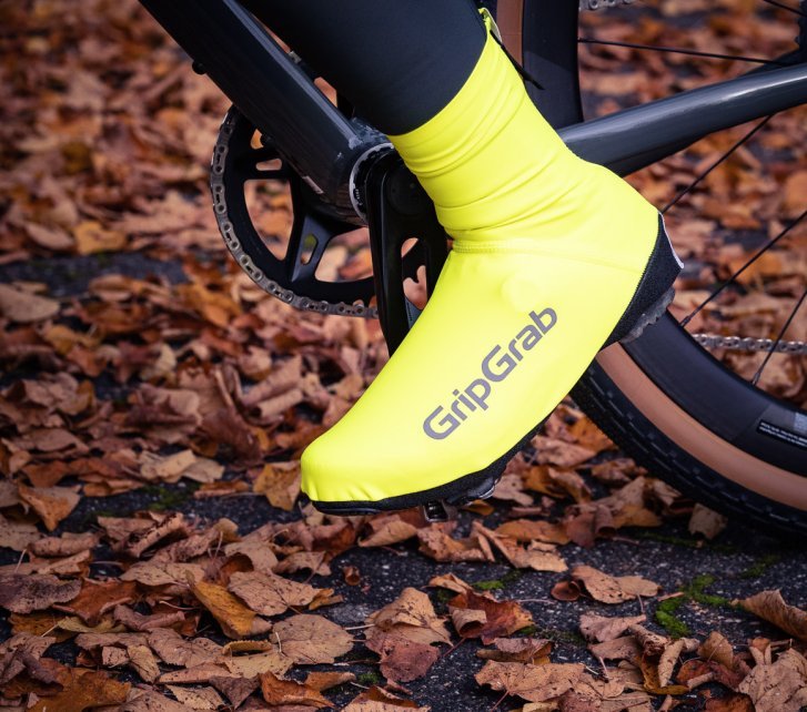 Un pie izquierdo dentro de un cubrecalzado ajustado, amarillo neón e impermeable, apoyado en un pedal de bicicleta. El suelo está cubierto de hojas otoñales rojizas.