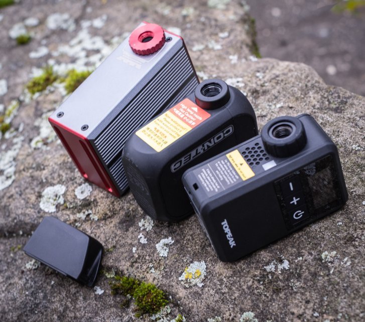 Drei kleine Akku-Pumpen liegen nebeneinander auf einem mit Flechten bewachsenen Stein. Darunter liegt ein SRAM AXS-Akku zum Größenvergleich 