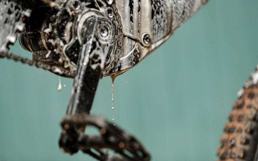 Nahaufnahme von Tretlager, Kettenblatt und Rahmen eines Fahrrads, die mit Reinigungsmittel eingesprüht sind. Schaum und verschmutztes Wasser tropfen vom Antrieb und Unterrohr herab.