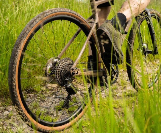 Roue arrière d’un vélo gravel sur chemin en gravier, pneu à flanc beige en action, cassette et dérailleur visibles dans un décor verdoyant.