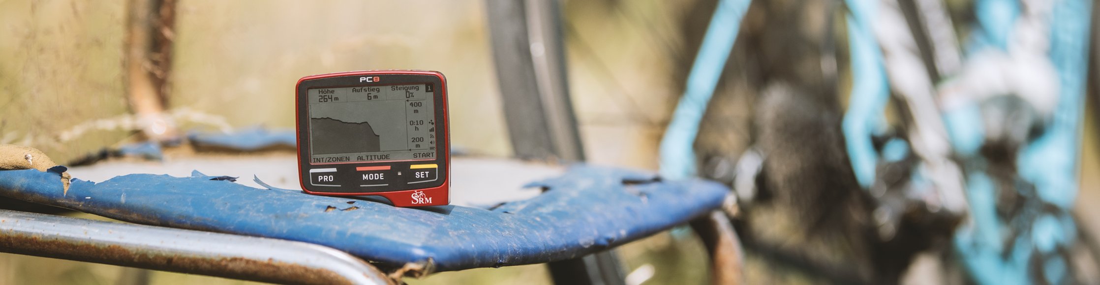 SRM Fahrradcomputer für Powermeter-Pedale steht auf einem alten Stuhl in der Landschaft, im Hintergrund ein himmelblaues Fahrrad