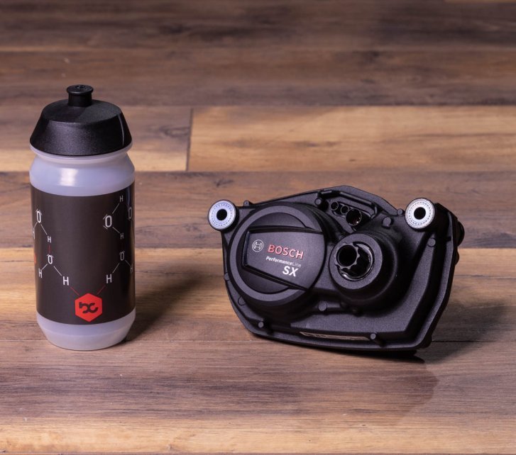A Bosch SX e-bike drive lies on a dark wooden floor. A bike water bottle stands next to it