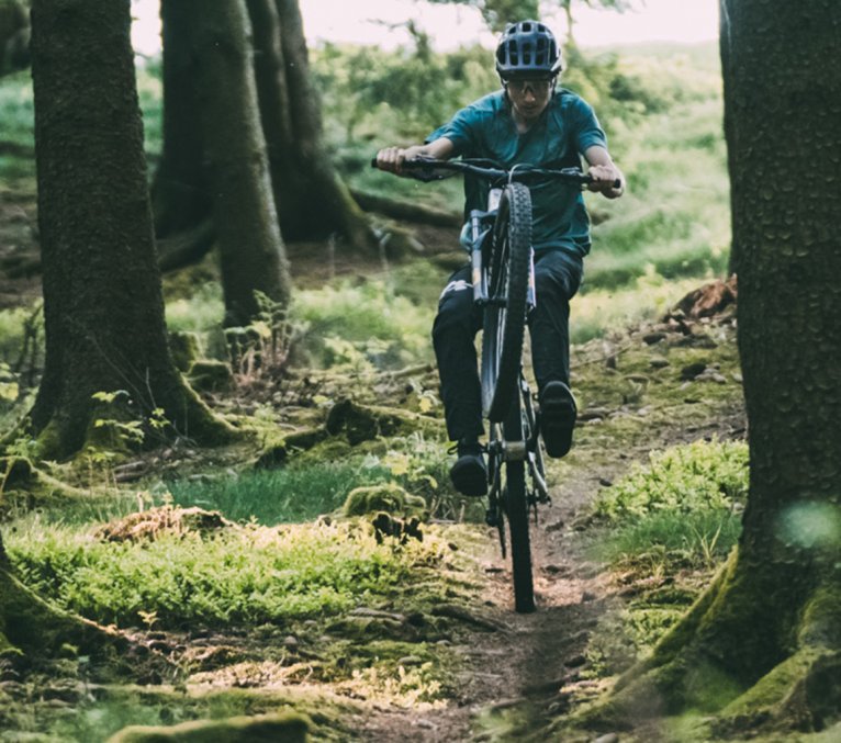 Christoph de bc fait un wheelie avec son VTT en descendant un trail de forêt.