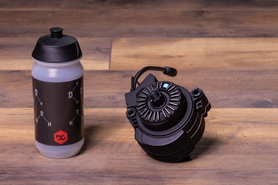A TQ HPR50 e-bike drive lies on a dark wooden floor. A bike water bottle stands next to it