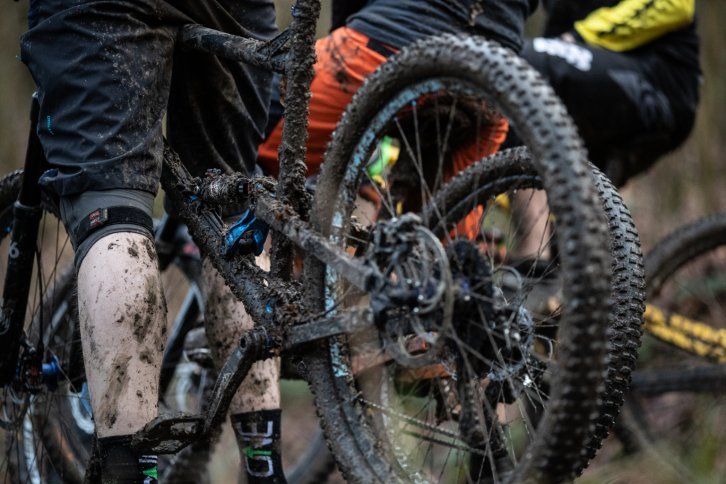 Eine sehr dreckige MTB-Hose und eine sehr dreckige Männerwade neben einem sehr dreckigen Mountainbike.