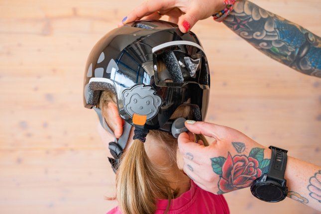 The tattooed hands of a mother are adjusting the fit of a bike helmet on a child's head with the size adjuster.