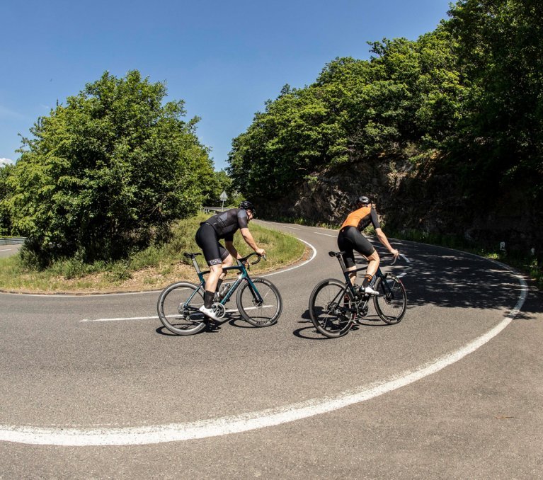 Zwei Rennradfahrer, beide auf Specialized Tarmac SL7 Bikes, passieren bergauf eine Serpentine.