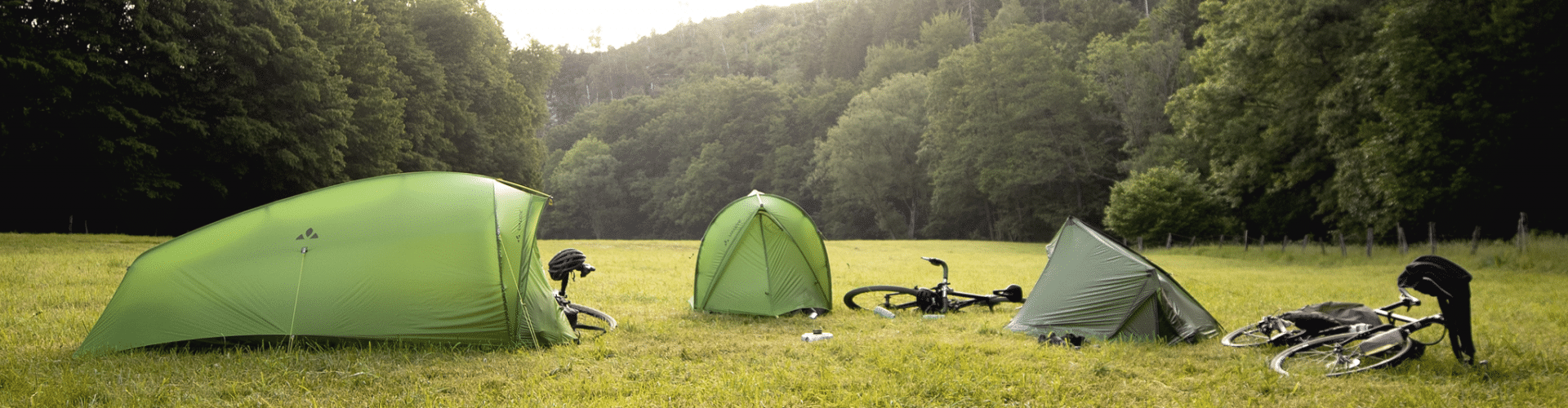 3 tentes VAUDE montées sur une prairie entourée d'arbres. Un vélo est posé à côté de chaque tente.