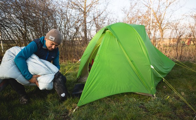 Marcel, de l'équipe marketing de bc, fourre son sac de couchage dans le sac de rangement correspondant. Il est agenouillé devant l'entrée de sa tente.